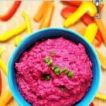 A bright pink beetroot hummus in a blue bowl surrounded by chopped peppers