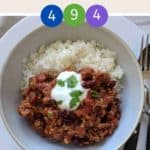 A white bowl filled with chili con carne & rice topped with yogurt and green herbs