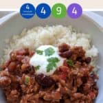 A white bowl filled with chili con carne & rice topped with yogurt and green herbs