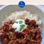 A white bowl filled with chili con carne & rice topped with yogurt and green herbs