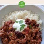 A white bowl filled with chili con carne & rice topped with yogurt and green herbs