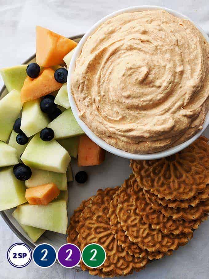 A white bowl of pumpkin fluff surrounded with fruits and cookies