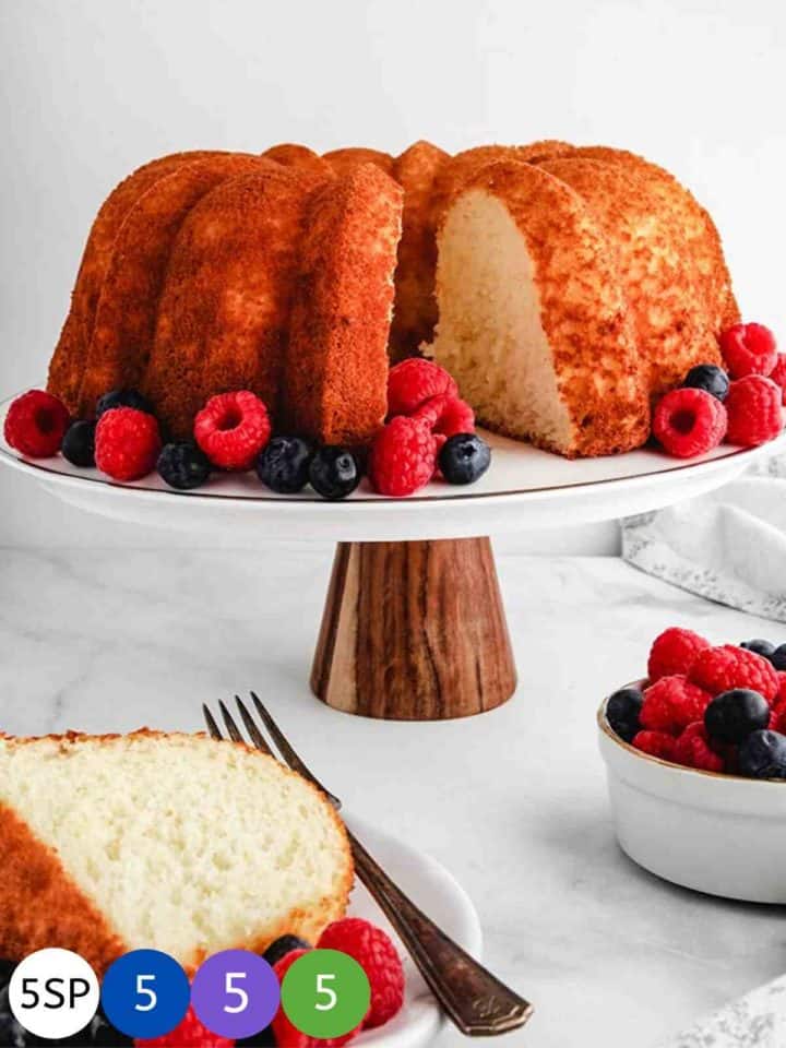 A Pineapple Angel Food cake on a white cake stand surrounded by raspberries and blueberries