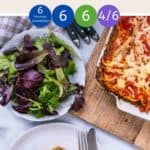 A dish of vegetable lasagna on a wooden board with a bowl of salad