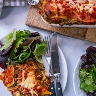 A white table with a dish of lasagna and a white plate with lasagna & salad