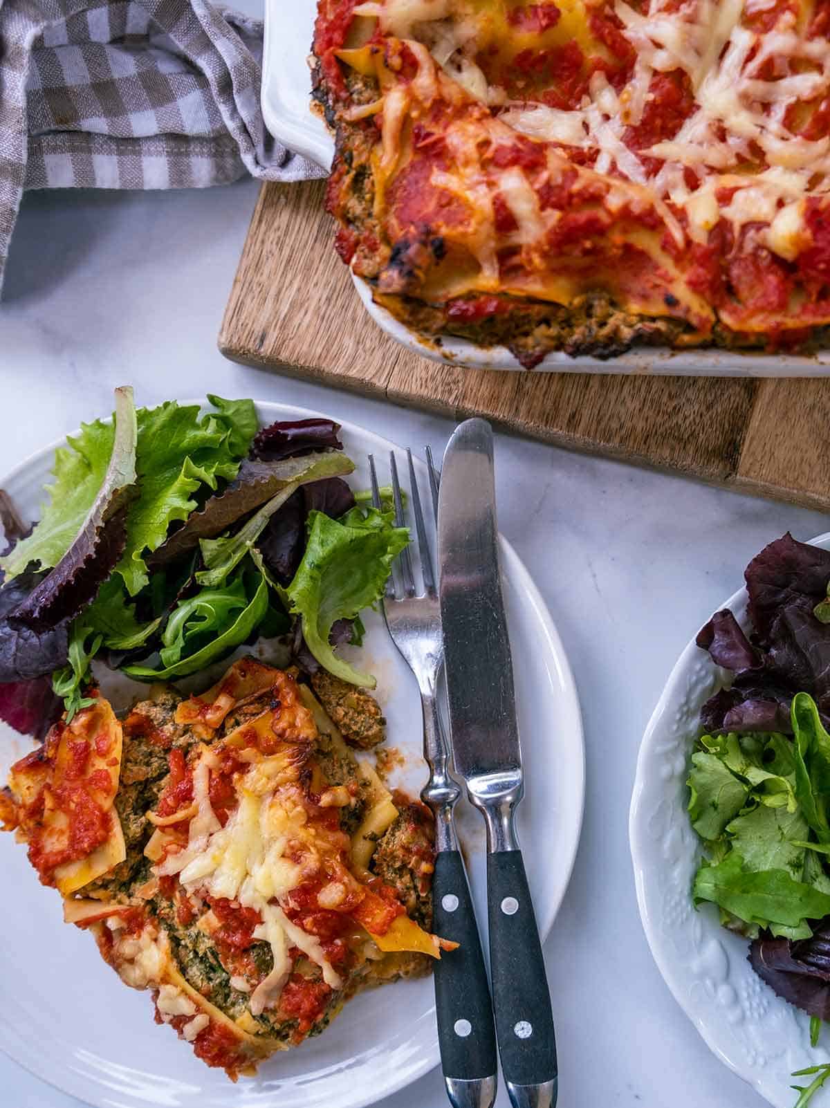 A white table with a dish of lasagna and a white plate with lasagna & salad