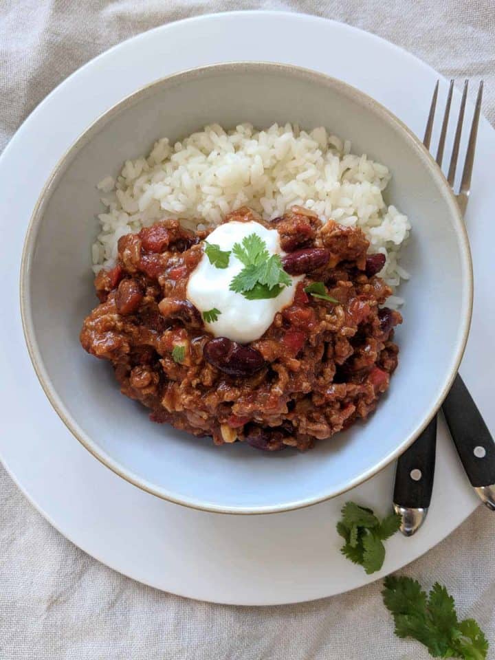 A white bowl of chili con carne and rice on a plate with a knife & fork and some herbs.