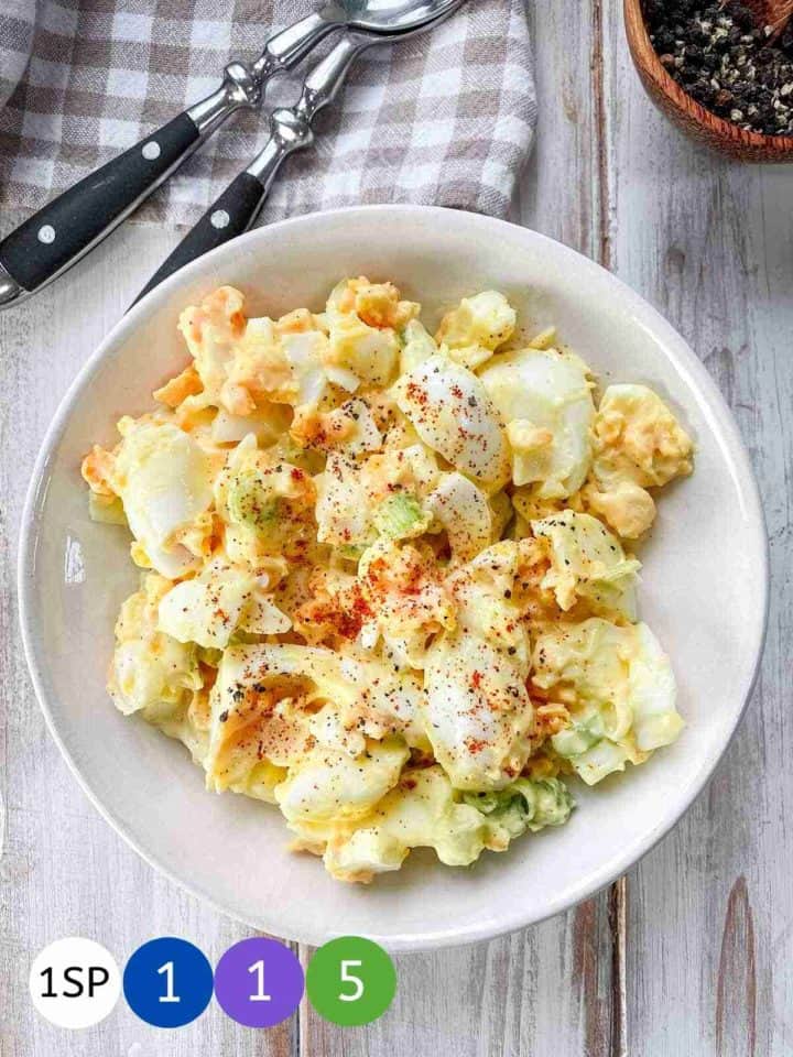 A white bowl of egg salad on a white wooden table