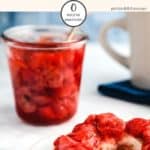 A glass jar full of strawberry jelly with a spoon sticking out and a bagel smothered with jelly infront on a white table.