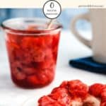 A glass jar full of strawberry jelly with a spoon sticking out and a bagel smothered with jelly infront on a white table.
