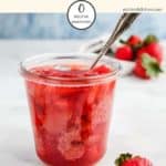 A glass jar full of strawberry jelly with a spoon sticking out on a white table.
