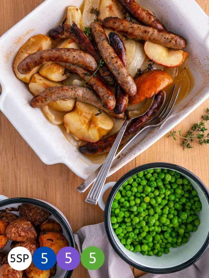 A white casserole dish full of sausages, onions and apples on a wooden table with a dish of garden peas and a dish of potatoes and 4 'counters' with WW point values.