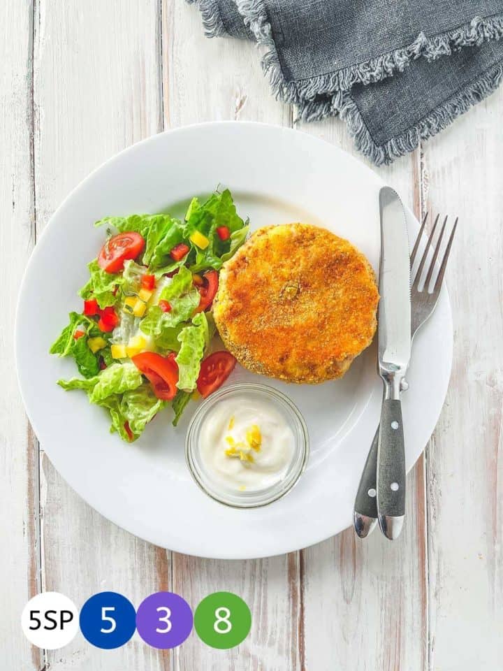 A white plate with a fishcake and some salad on a white wooden table.